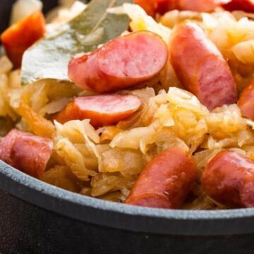 A close-up of a black pot filled with cooked sauerkraut and sliced sausages. The dish is garnished with bay leaves, and the ingredients appear well-mixed and cooked together.