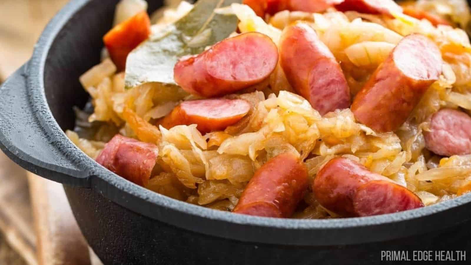 A close-up of a black pot filled with cooked sauerkraut and sliced sausages. The dish is garnished with bay leaves, and the ingredients appear well-mixed and cooked together.