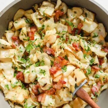 A skillet filled with cooked cabbage and bacon, garnished with chopped parsley, offers a delicious money-saving recipe. A wooden spoon rests inside the skillet, ready to serve bite-sized cabbage pieces mixed with crispy bacon chunks—perfect for tight weeks without skimping on flavor.