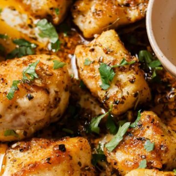 Close-up of golden-brown gnocchi pieces garnished with herbs and spices, drizzled with a sauce or oil. A small bowl with a light-colored dipping sauce is visible on the side.