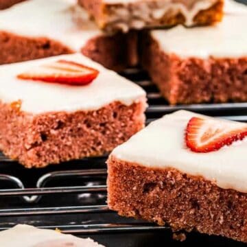 Close-up of sliced strawberry cake squares with white icing, each topped with a slice of strawberry, placed on a wire rack. Whole strawberries are scattered around the rack.