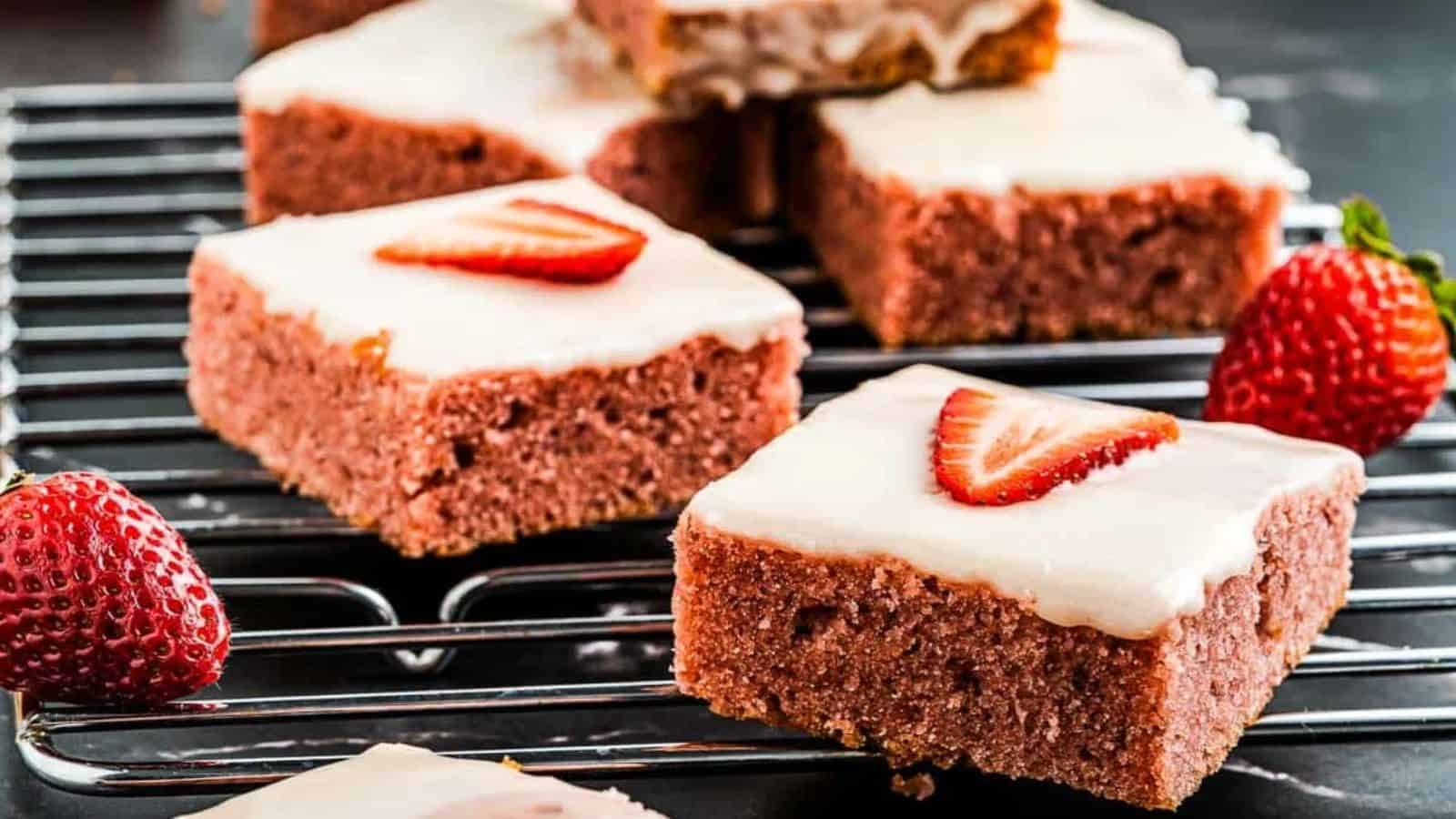 The image showcases moist pink dessert bars topped with a creamy glaze and fresh strawberry slices, arranged on a cooling rack with scattered strawberries.