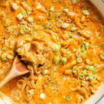 A pan filled with a creamy, orange-colored sauce, topped with chopped green onions and pepper. A wooden spoon is stirring the mixture. The background includes a small dish of additional green onions.
