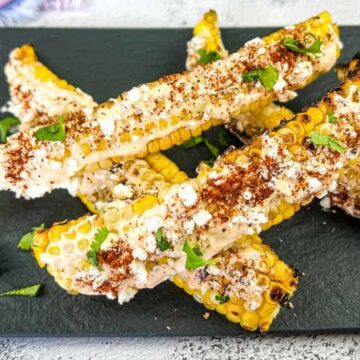 Grilled corn ribs on a slate plate, topped with crumbled cheese, herbs, and spices, accompanied by lime wedges. This show-stealing side dish is perfect for any dinner gathering.