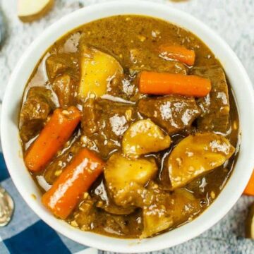 A bowl of beef stew is filled with chunks of beef, carrots, and potatoes in a rich brown broth. A silver spoon lies to the side, and a blue-and-white checked napkin is partially visible next to the bowl.