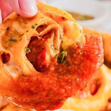 A hand holds a baked pizza roll filled with melted cheese, pepperoni, and tomato sauce. The roll is topped with herbs, and the background shows more of the same rolls slightly out of focus.