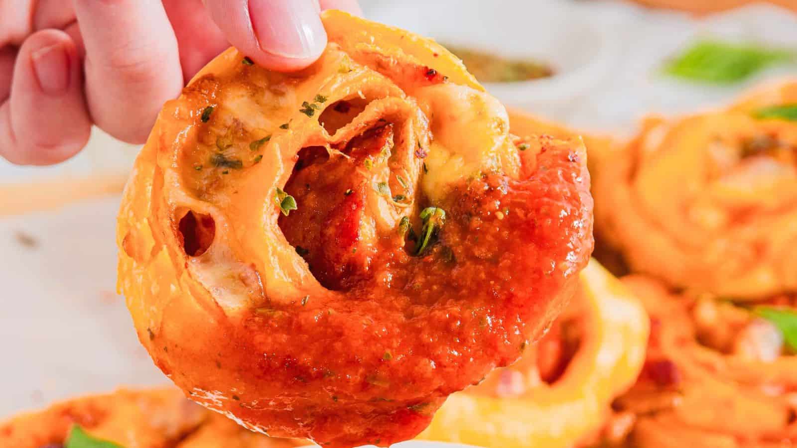 A hand dips a slice of pizza roll with visible cheese and pepperoni into a bowl of red marinara sauce. A basil leaf is partially visible in the background, resting on a wooden surface.