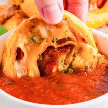 A hand dips a cheesy pinwheel into a bowl of red marinara sauce. The pinwheels are golden-brown and filled with melted cheese and pieces of pepperoni. A basil leaf is visible in the background on a wooden board.
