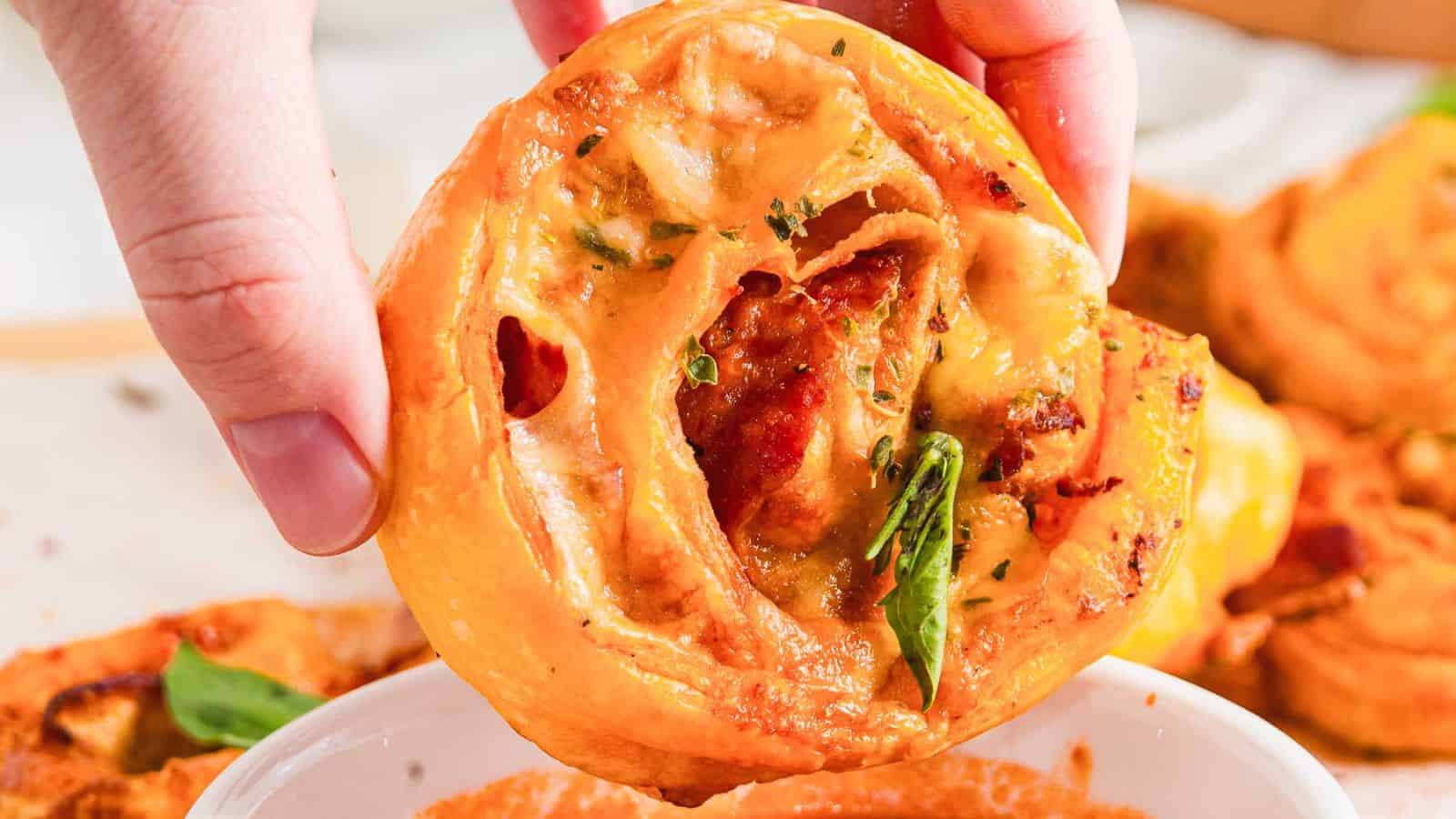 A hand dips a slice of pizza roll with visible cheese and pepperoni into a bowl of red marinara sauce. A basil leaf is partially visible in the background, resting on a wooden surface.