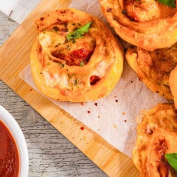 A wooden board holds several pizza rolls topped with basil leaves, set on parchment paper. A small bowl of marinara sauce is on the left. The table surface is light gray with visible grain patterns.