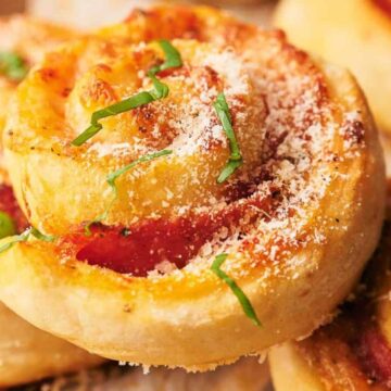 Close-up of several pizza rolls topped with grated cheese and garnished with thinly sliced green herbs. The rolls have a golden, crispy appearance and a visible tomato and cheese filling spiraled inside.