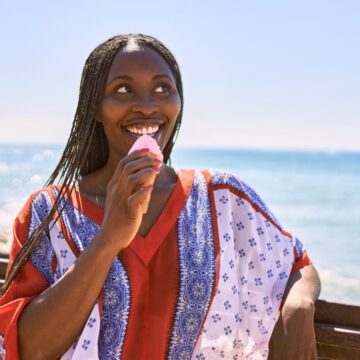 A person enjoying an ice cream stands near a wooden railing by the sea on a sunny day. They are wearing a brightly patterned top and smiling while looking to the side. The ocean and clear blue sky are visible in the background.