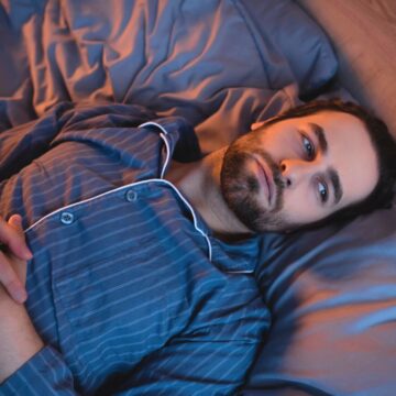 A person with dark hair and a beard lies awake in bed, wearing striped blue pajamas. They are gazing upward with hands resting on their chest. The bedding is a matching blue color, and faint light casts soft shadows in the room.