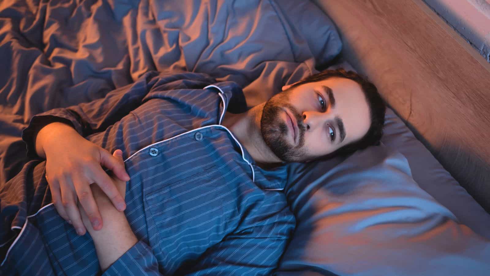 A person with dark hair and a beard lies awake in bed, wearing striped blue pajamas. They are gazing upward with hands resting on their chest. The bedding is a matching blue color, and faint light casts soft shadows in the room.