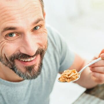 A man with a beard and short hair smiles while holding a spoonful of cereal close to his mouth. He is sitting at a table with leafy greens in the background, wearing a light gray shirt.