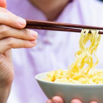 A person is holding chopsticks and lifting a portion of cooked yellow noodles from a white bowl. The background is blurred, and the person is wearing a light-colored top.