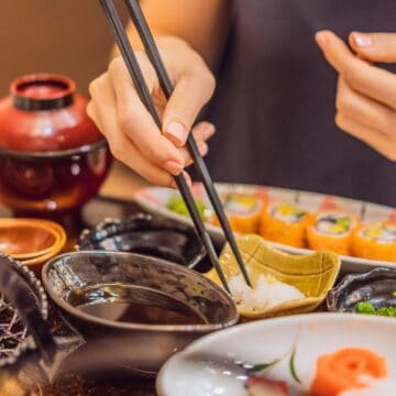 A person is using chopsticks to pick up food from a small bowl. The table has a variety of dishes, including sushi rolls, tempura, and dipping sauces. A red bowl is visible in the background. The setting appears to be a Japanese meal.
