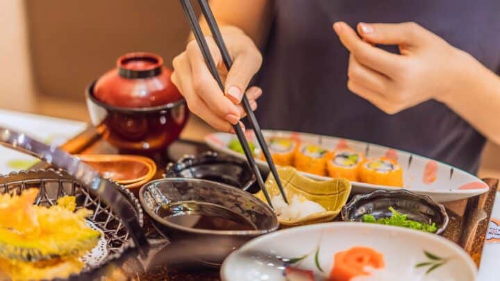 A person is using chopsticks to pick up food from a small bowl. The table has a variety of dishes, including sushi rolls, tempura, and dipping sauces. A red bowl is visible in the background. The setting appears to be a Japanese meal.