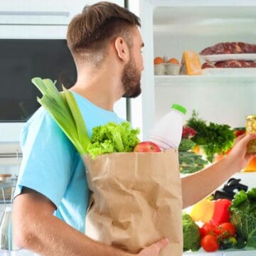 A man with short hair, wearing a light blue shirt, stands near an open refrigerator. He holds a paper grocery bag filled with vegetables and a milk bottle, while reaching inside the fridge. The kitchen has a white microwave and various kitchen items.