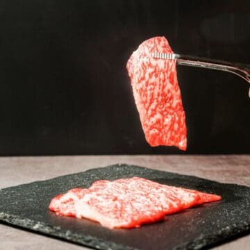 A hand holds a piece of marbled raw meat with tongs above a slate serving board. Two more slices of similar meat are placed on the board. The background is black, and the surface is a gray textured material.