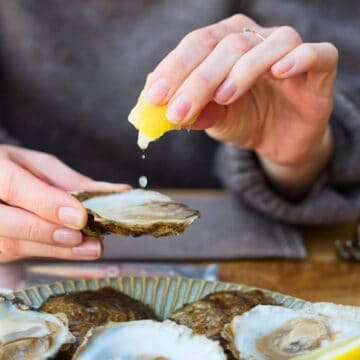 A person squeezing a lemon wedge onto shucked oysters on a plate. The oysters are arranged on a wooden table, and a folded napkin with cutlery is visible in the background.