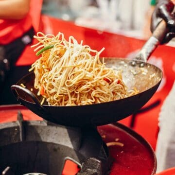 A person is preparing stir-fried noodles in a wok. The noodles are being tossed in the air above the wok, with visible vegetables like bean sprouts and green onions. The background has a blurry crowd and red elements.