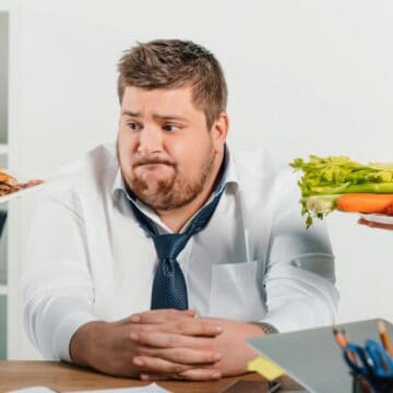 A person sits at a desk with a conflicted expression. In front of them, one hand offers a plate of fries and a burger while another hand presents a plate of vegetables, including carrots, celery, and cherry tomatoes. Office supplies are on the desk.