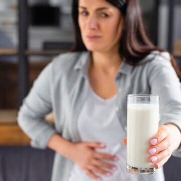 A woman stands in a room holding a glass of milk in her outstretched hand, while her other hand is placed on her stomach. She appears to be experiencing discomfort. Shelves with various items are in the background.