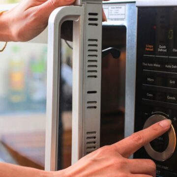 A person wearing a bracelet opens a microwave door with one hand while adjusting the settings with the other. The microwave control panel has options for quick defrost and clock functions.