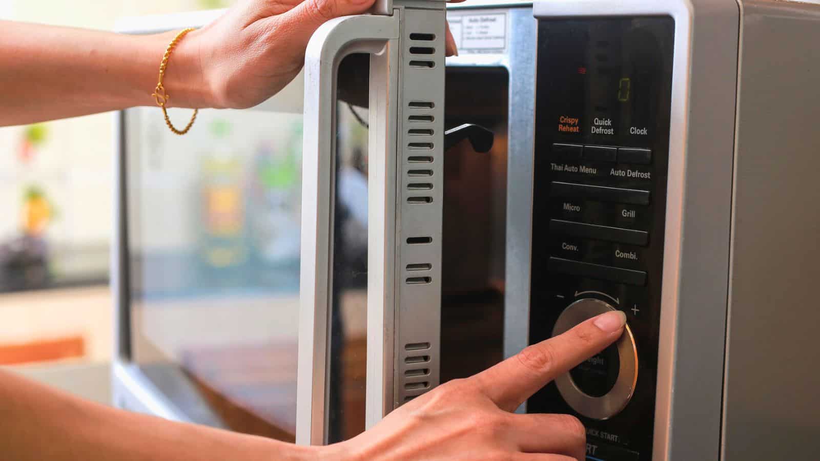 A person wearing a bracelet opens a microwave door with one hand while adjusting the settings with the other. The microwave control panel has options for quick defrost and clock functions.