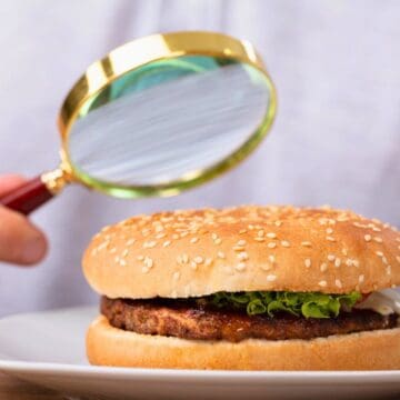 A person uses a magnifying glass to closely examine a hamburger on a white plate. The burger has a sesame seed bun, lettuce, and a visible patty. The background is blurred, focusing attention on the burger and magnifying glass.