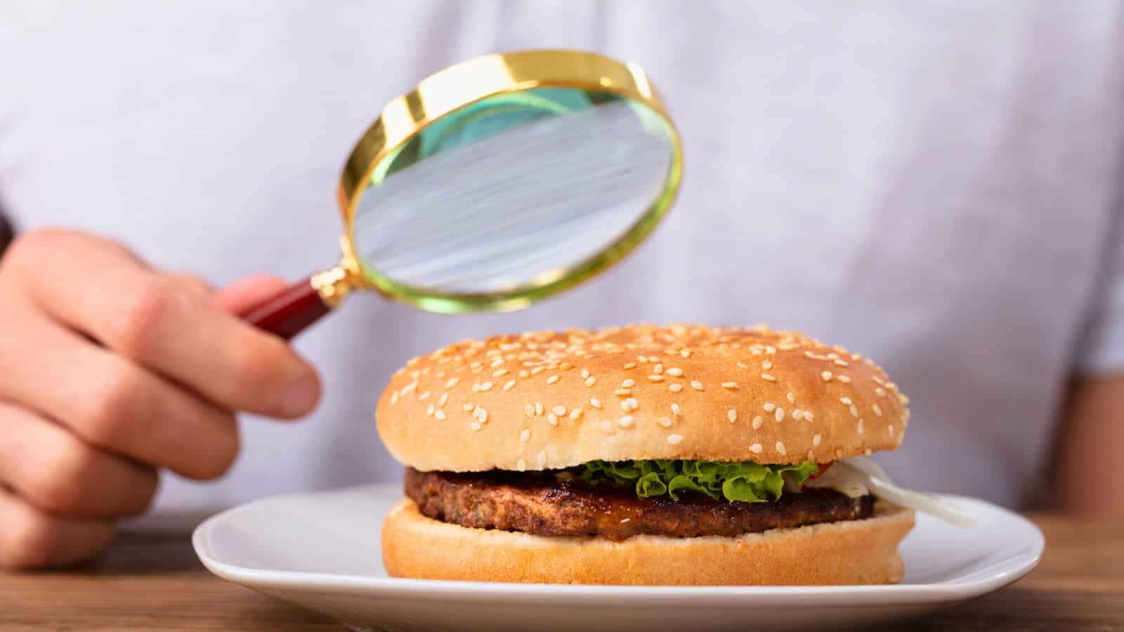 A person uses a magnifying glass to closely examine a hamburger on a white plate. The burger has a sesame seed bun, lettuce, and a visible patty. The background is blurred, focusing attention on the burger and magnifying glass.