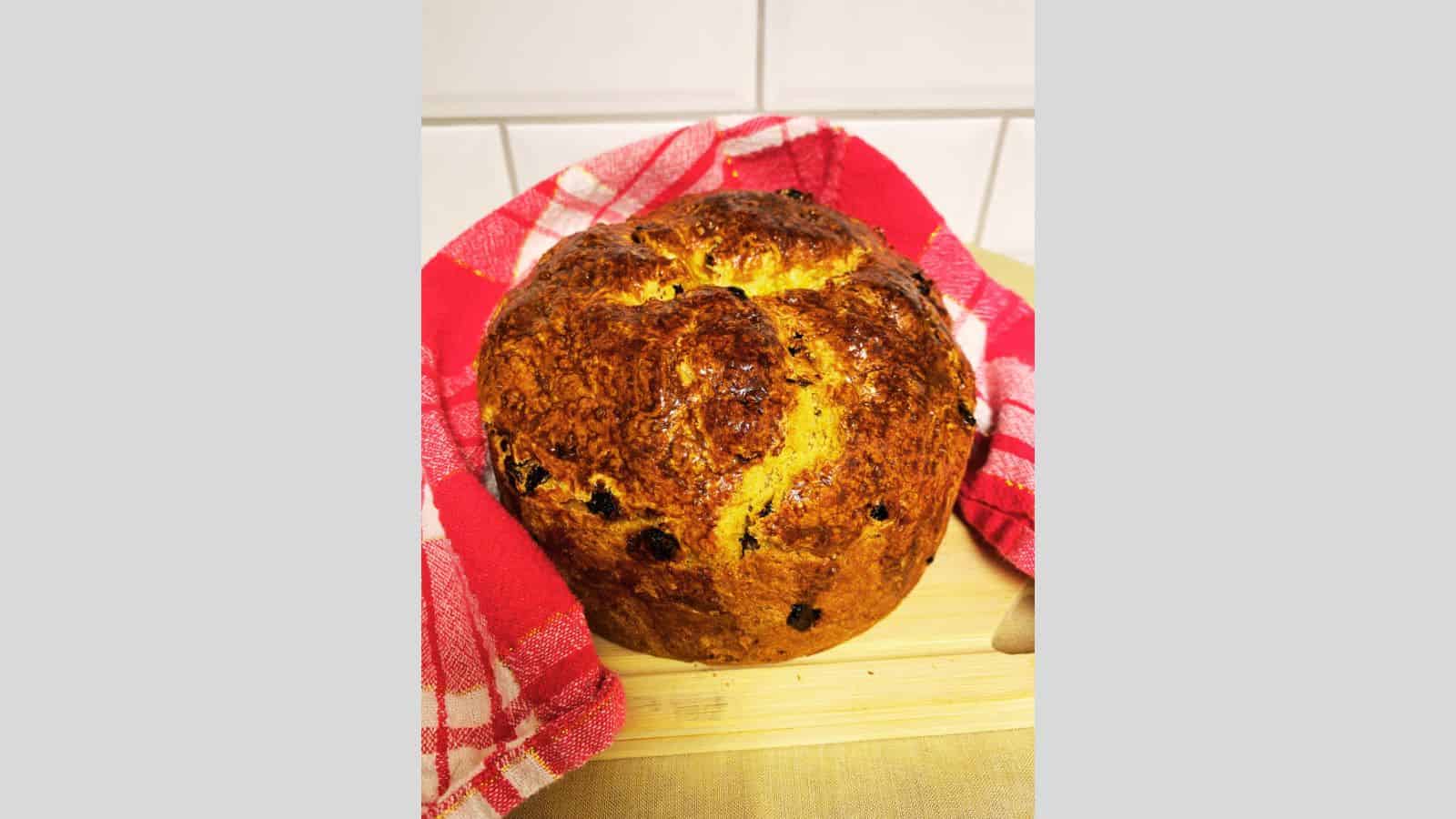 A freshly baked loaf of bread with a golden crust sits on a wooden cutting board. It is partially wrapped in a red and white checkered cloth. The bread has raisins embedded in its surface.