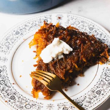 A slice of cake with a caramelized topping and a dollop of cream sits on a decorative plate. A fork rests on the plate. Two cinnamon sticks are nearby.