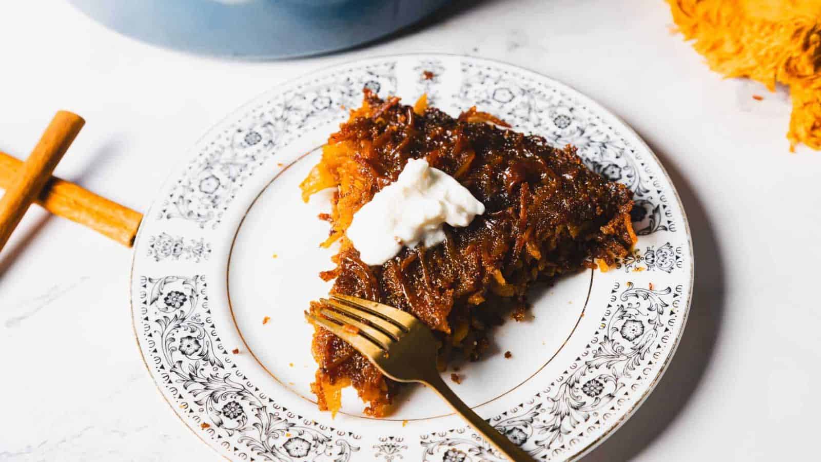 A slice of cake with a caramelized topping and a dollop of cream sits on a decorative plate. A fork rests on the plate. Two cinnamon sticks are nearby.