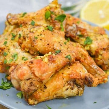 A plate of cooked chicken wings garnished with herbs is displayed. A sliced lemon is in the background on a gray napkin.