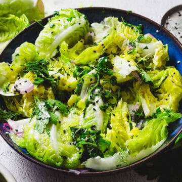 A bowl of fresh salad containing leafy greens, herbs, and sliced shallots. The salad is garnished with black sesame seeds and drizzled with dressing. The dish is served in a dark blue bowl on a white surface.