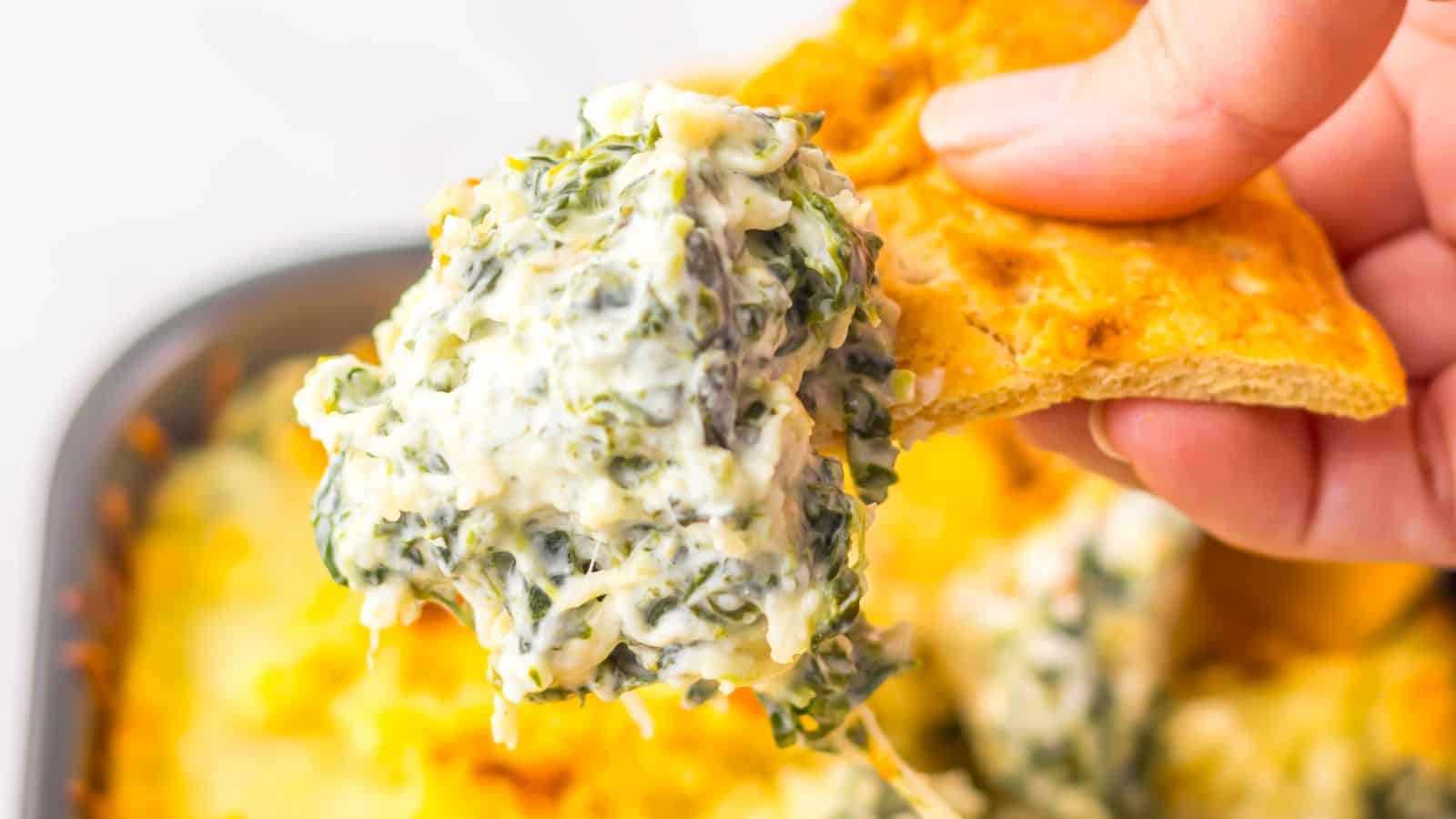 A close-up of spinach artichoke dip on a cracker held by a hand. The dip is creamy with visible green spinach and bits of artichoke. The background shows a baking dish filled with the dip.