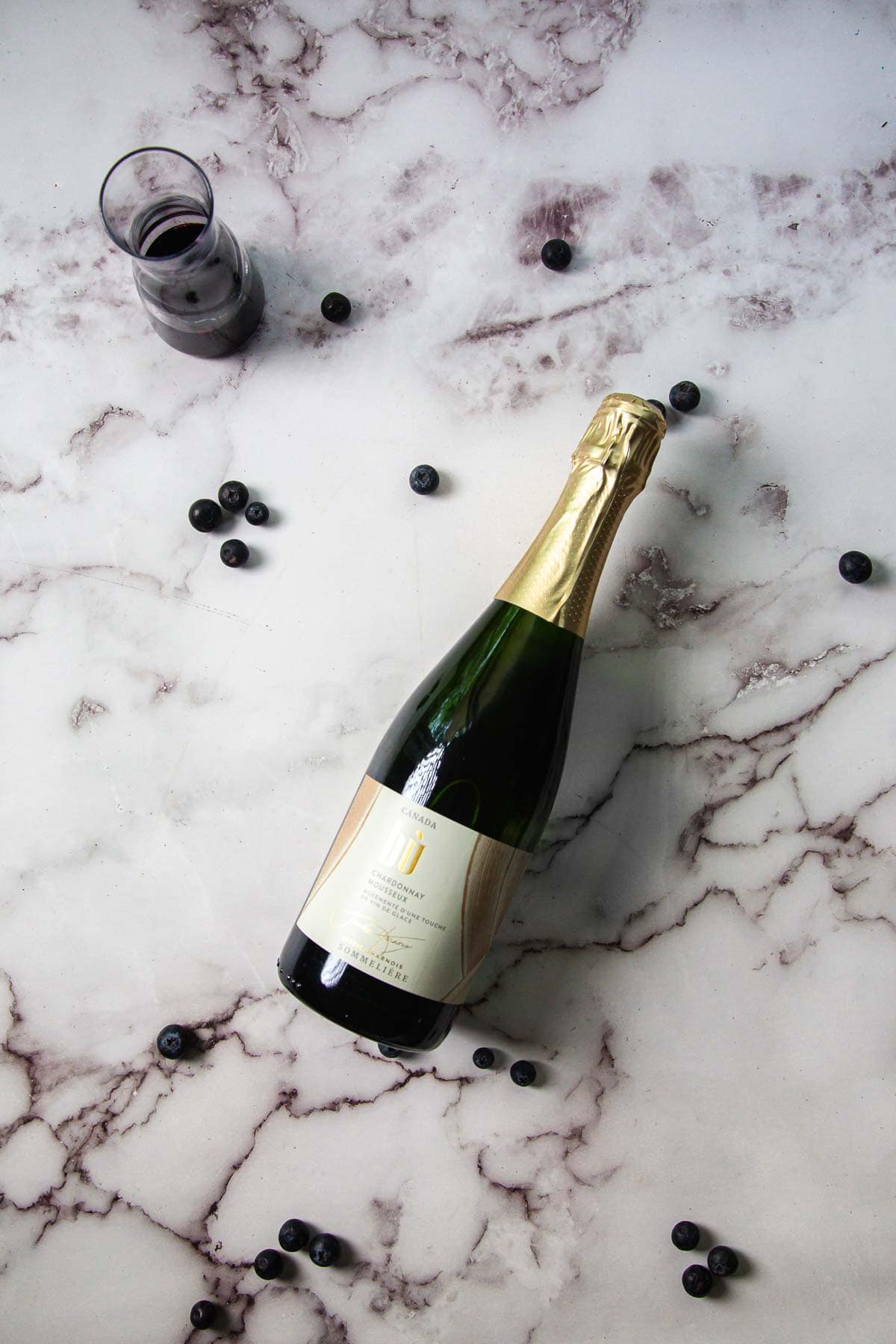 A bottle of champagne lies on a marble surface surrounded by scattered blueberries, reminiscent of the elegant Kir Royale cocktail. A half-filled glass is visible in the upper left corner, hinting at a celebration waiting to unfold.