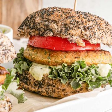 A plant-based burger is placed on a white rectangular dish. It has a poppy seed bun and is filled with lettuce, a vegan patty, sliced tomato, and a green creamy sauce. Another similar burger is partially visible next to it.