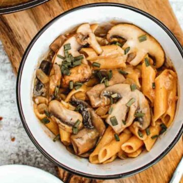 A white bowl filled with penne pasta and sliced mushrooms, garnished with chopped herbs. The bowl is on a wooden surface, accompanied by a fork. The background features a textured surface with scattered small grains.