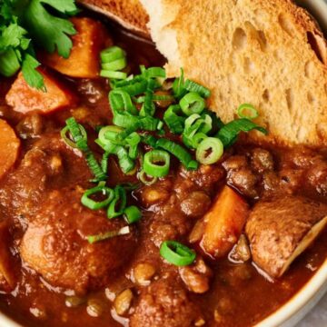 A bowl of hearty stew containing lentils, potatoes, and carrots, garnished with chopped green onions and a sprig of cilantro. A slice of crusty bread is placed on the rim of the bowl.