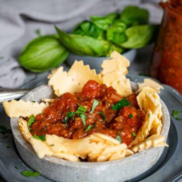 A gray bowl with broken pasta pieces surrounds a rich, tomato-based sauce—perfect for simple dinners when you're feeling lazy. Fresh basil leaves add a fragrant touch in the background, alongside a glass container of extra sauce. A vintage silver fork rests beside the bowl.