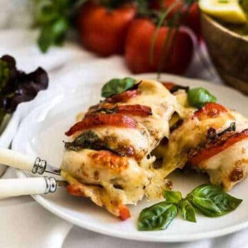 A plate of stuffed chicken breast with cheese and tomato, garnished with basil leaves. In the background, there are fresh tomatoes, a bowl of salad, and leafy greens. Two ornate forks are placed on the plate.