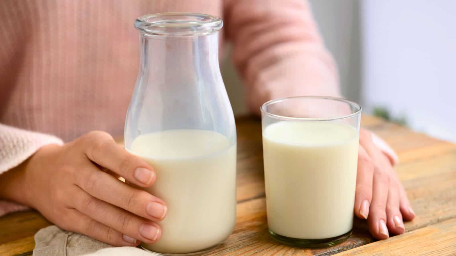 A person in a pink sweater is holding a glass jug of milk on a wooden table. A filled glass of milk is placed next to the jug. The person’s hands are partially visible, and the background is softly blurred.
