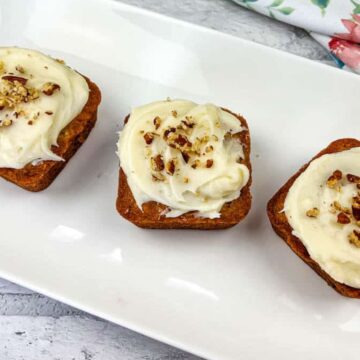 Three square carrot cake bites topped with cream cheese frosting and sprinkled with nuts are arranged on a white rectangular plate. A floral-patterned cloth is partially visible in the background on a light stone surface.