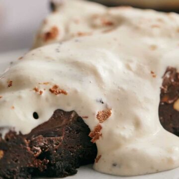 A slice of chocolate cake topped with a generous layer of white cream on a white plate. The cake has a rich, dense texture with visible nuts embedded in it. The cream appears smooth and slightly drips over the edges of the cake.