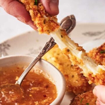 A person pulls apart a breaded and fried mozzarella stick, showing melted cheese stretching between two halves. The stick is being dipped into a bowl of sauce on a patterned plate.