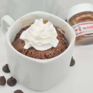 A chocolate mug cake topped with whipped cream sits on a white plate, sweetened with a few chocolate chips. A jar of Nutella lingers in the background, while a spoon rests beside the mug, making it an ideal treat for Valentine's Day desserts.