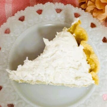 A slice of cream pie rests on a decorative white plate with a scalloped edge, set atop a pink and white striped cloth. A peach-colored flower adds charm, making this scene reminiscent of classic boomer dessert recipes that sweeten your day with nostalgia.